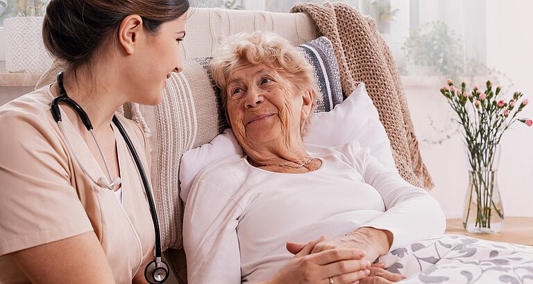 Senorin liegt im Bett, fürsorgliche Doktorin hält ihre Hand